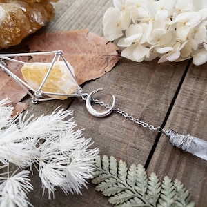 May include: A silver necklace with a geometric crystal cage pendant, a crescent moon charm, and a clear quartz crystal wrapped in wire. The necklace is displayed on a wooden surface with dried flowers and leaves.