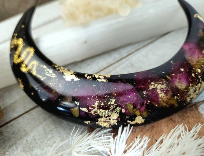 May include: A black crescent moon-shaped resin ornament with embedded dried flowers and gold leaf accents.