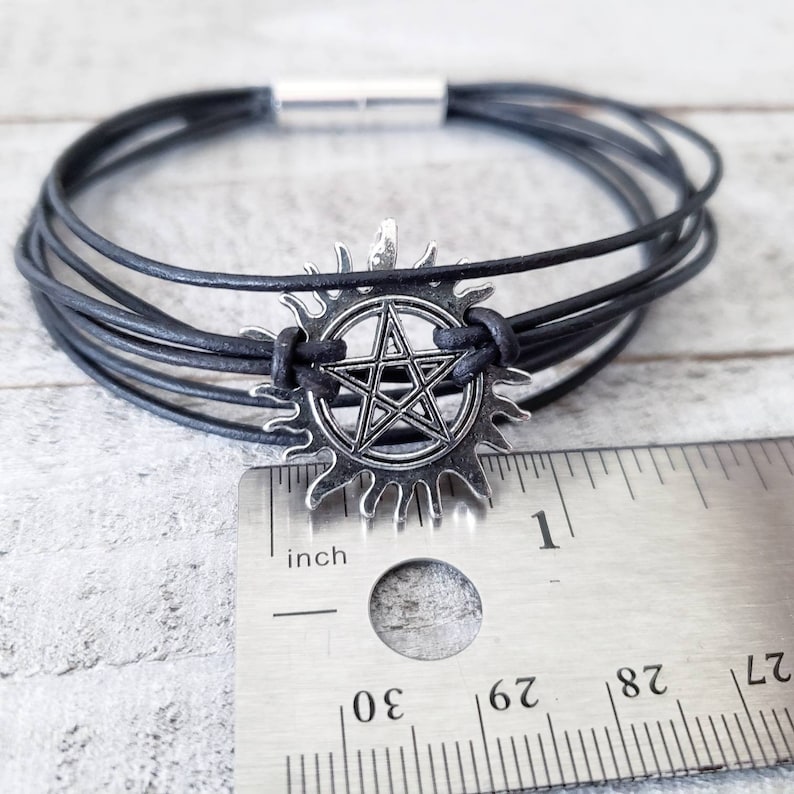 May include: Black leather bracelet with a silver pentagram charm. The charm is a five-pointed star inside a sunburst design. The bracelet is measured against a ruler, showing it is about 1 inch in diameter.