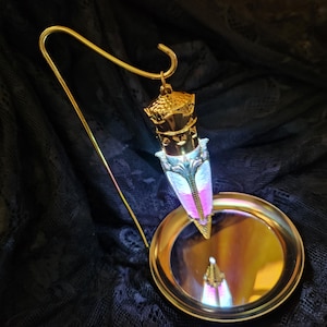 May include: A gold-colored, ornate perfume bottle with a long, curved stem and a small, round base. The bottle has a decorative top and a central section with pink and blue accents. The base reflects the bottle's image.