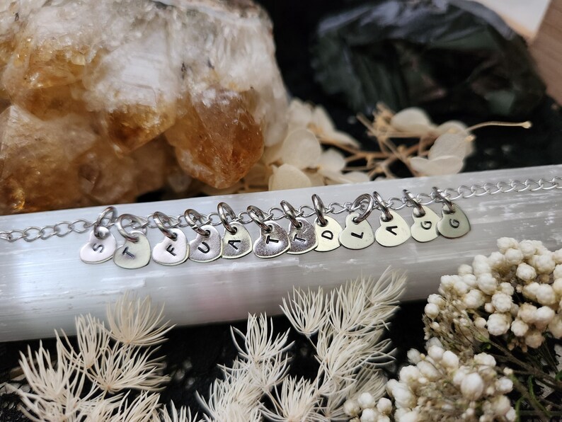 May include: A silver chain necklace with silver heart charms, each charm has a letter stamped on it, spelling out the word "STUART".