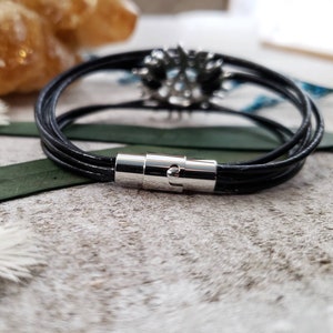 May include: A black leather bracelet with a silver magnetic clasp. The bracelet is made of multiple strands of leather and has a simple, elegant design.