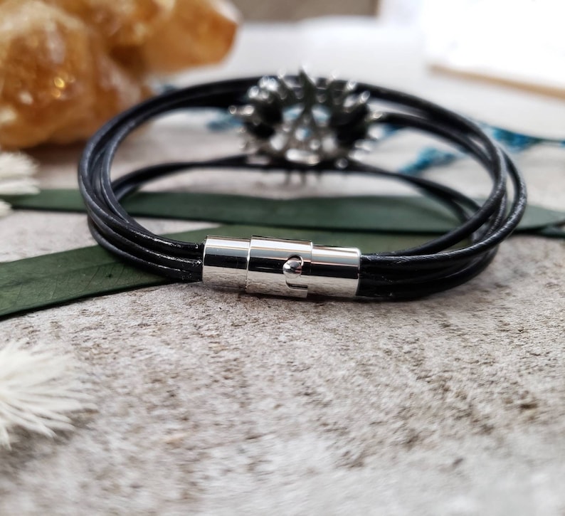 May include: A black leather bracelet with a silver magnetic clasp. The bracelet is made of multiple strands of leather and has a simple, elegant design.