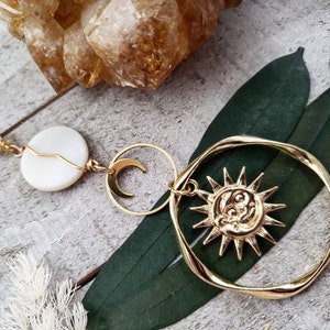 May include: Gold sun and moon charm necklace with a white shell bead and a clear crystal bead. The necklace is on a wooden surface with green leaves.