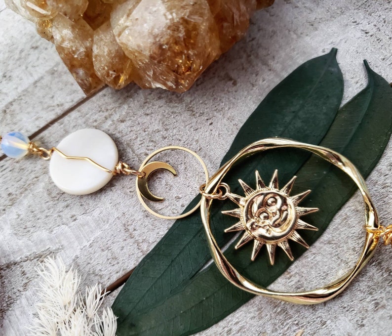 May include: Gold sun and moon charm necklace with a white shell bead and a clear crystal bead. The necklace is on a wooden surface with green leaves.