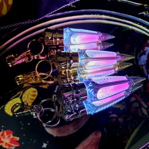 May include: Keychains with a metallic finish and a decorative, stylized design. The keychains have a gold, silver, and iridescent finish, with a pink and blue glow emanating from the center. The keychains are arranged on a dark, reflective surface.