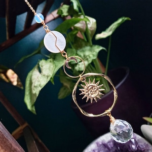 May include: Gold sun and moon charm with a white shell and clear crystal pendant. The charm hangs from a gold chain.