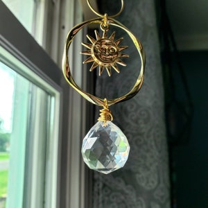 May include: A gold sun with a face charm hangs from a crystal prism. The sun charm is attached to a gold ring and the crystal prism is attached to a gold chain.
