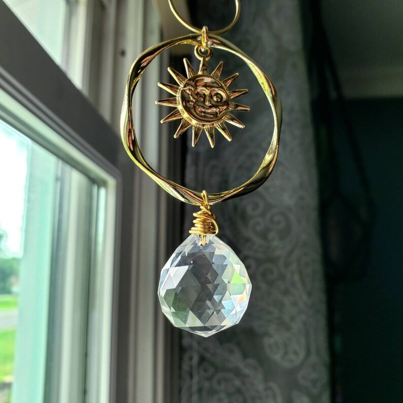 May include: A gold sun with a face charm hangs from a crystal prism. The sun charm is attached to a gold ring and the crystal prism is attached to a gold chain.