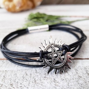 May include: Black leather bracelet with a silver charm featuring a five-pointed star inside a circle with sun rays.