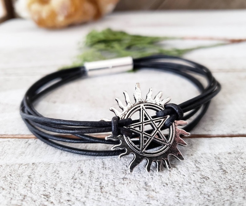 May include: Black leather bracelet with a silver charm featuring a five-pointed star inside a circle with sun rays.