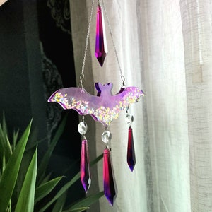May include: A purple and iridescent bat-shaped wind chime with crystal drops hanging from it. The bat is decorated with glitter and the crystals are a deep purple color.