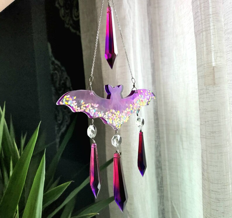May include: A purple and iridescent bat-shaped wind chime with crystal drops hanging from it. The bat is decorated with glitter and the crystals are a deep purple color.
