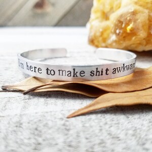 May include: A silver cuff bracelet with the stamped phrase "I'm here to make shit awkward." The bracelet is displayed on a rustic wooden surface with dried leaves and a blurred yellow crystal in the background.