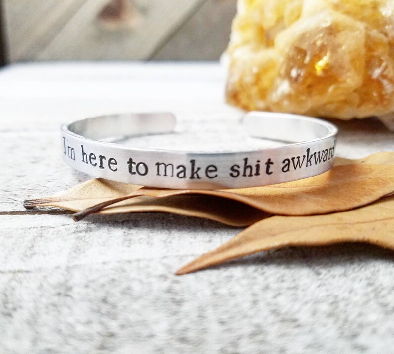 May include: A silver cuff bracelet with the stamped phrase "I'm here to make shit awkward." The bracelet is displayed on a rustic wooden surface with dried leaves and a blurred yellow crystal in the background.
