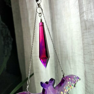 May include: A purple and iridescent bat-shaped resin ornament with a dangling crystal prism. The prism is a deep magenta color and hangs from a silver chain.