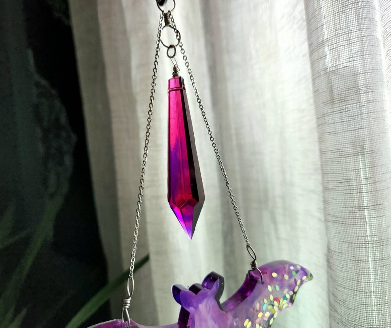 May include: A purple and iridescent bat-shaped resin ornament with a dangling crystal prism. The prism is a deep magenta color and hangs from a silver chain.