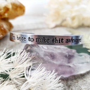 May include: A silver cuff bracelet with the stamped text "I'm here to make shit awkward." The bracelet is displayed on a purple crystal with white accents.