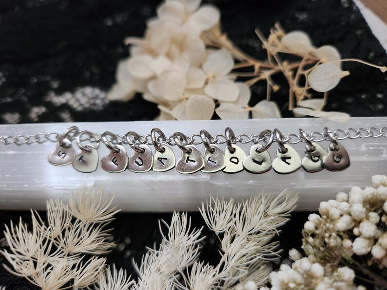 May include: A silver chain necklace with heart-shaped charms, each charm has a letter stamped on it, spelling out the word "TRUST".