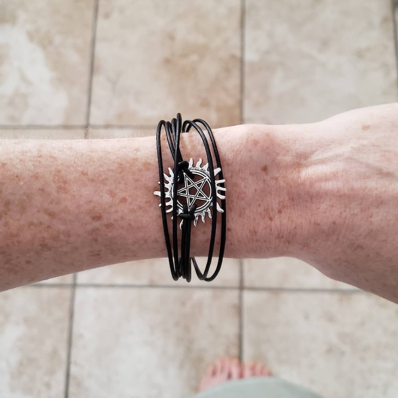 May include: A black leather bracelet with a silver charm featuring a pentagram inside a circle.