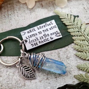 May include: A silver keychain with a charm that says "walk barefoot, listen to the wind, drink in the moon, be magic". The charm is attached to a blue crystal wrapped in silver wire and a silver leaf charm.