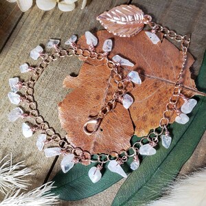 May include: A rose gold chain bracelet with a leaf charm and multiple white moonstone charms hanging from it. The bracelet is lying on a brown leaf.