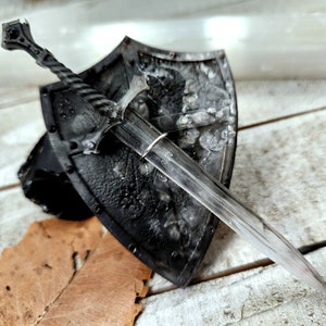 Peut inclure: Un bouclier noir avec une épée argentée attachée. Le bouclier a une surface texturée et l'épée a une lame argentée et un manche noir.