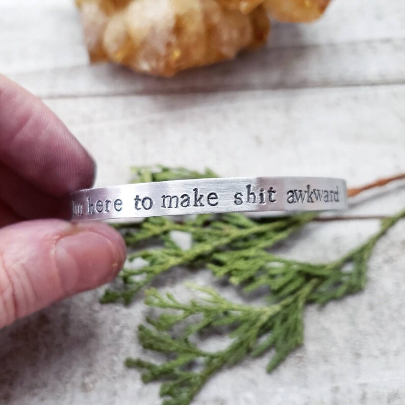 May include: A silver-colored metal cuff bracelet with the stamped text "I'm here to make shit awkward." The bracelet is held against a background of greenery and a light-colored surface.