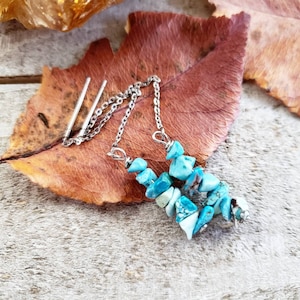 May include: A pair of turquoise gemstone earrings with a silver chain connecting them. The earrings are on a brown leaf.