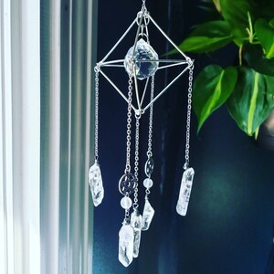 May include: A silver geometric crystal mobile with a large crystal prism in the center and smaller crystal points hanging from the bottom. The mobile is suspended from a silver chain.