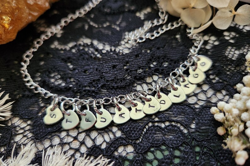 May include: A silver chain necklace with a series of small silver heart charms, each with a letter stamped on it, spelling out the word "STUFFING".