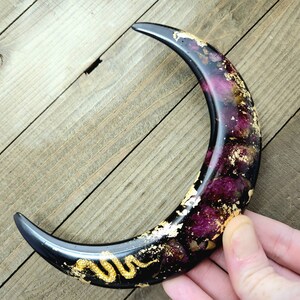 May include: A black crescent moon resin ornament with gold leaf and dried flowers embedded inside. A gold snake is curled on the bottom of the moon.