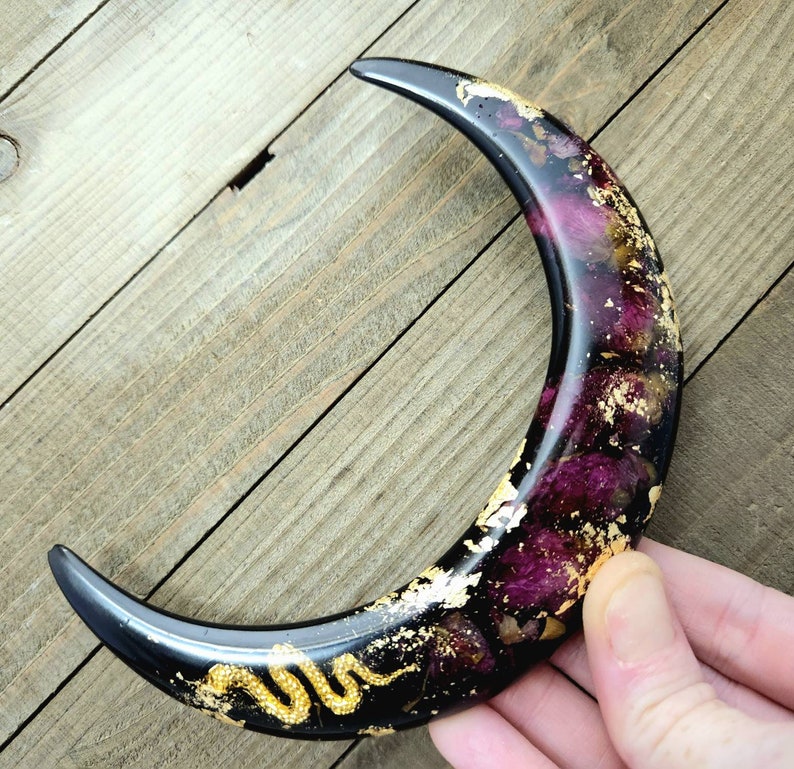 May include: A black crescent moon resin ornament with gold leaf and dried flowers embedded inside. A gold snake is curled on the bottom of the moon.