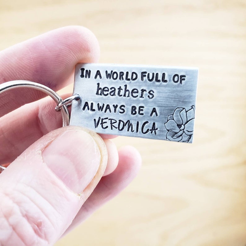 Pu&ograve; includere: Un portachiavi d'argento con un design a fiore e il testo "In un mondo pieno di erica, sii sempre una Veronica".