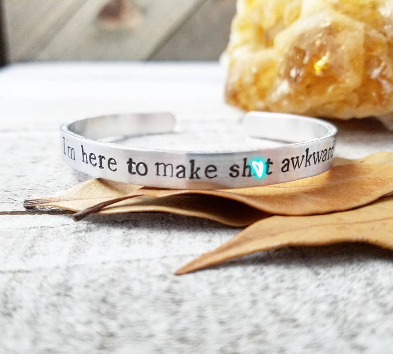 May include: A silver cuff bracelet with the stamped text "I'm here to make sh!t awkward" and a small turquoise heart symbol. The bracelet is displayed on a white and gray surface with dried leaves.