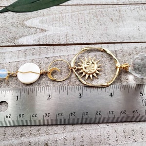 May include: Gold sun and moon charm necklace with a clear crystal pendant and white shell bead. The necklace is measured on a ruler with inches.