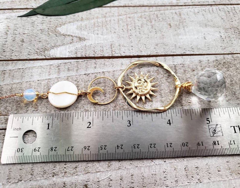 May include: Gold sun and moon charm necklace with a clear crystal pendant and white shell bead. The necklace is measured on a ruler with inches.