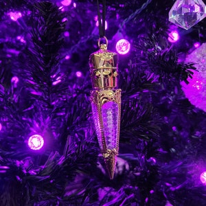 May include: A gold-colored Christmas ornament shaped like a bottle with a clear glass section filled with glitter. The ornament is hanging on a black artificial Christmas tree with purple lights.