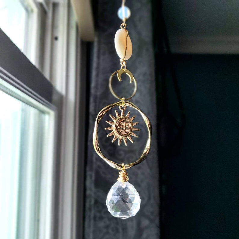 May include: A gold sun and moon charm hangs from a crystal prism. The charm is attached to a white shell and a gold chain.