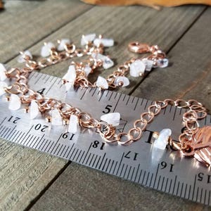 May include: A rose gold chain bracelet with a feather charm and white gemstone accents. The bracelet is measured against a ruler.