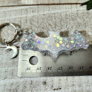 Pode incluir: Um chaveiro de morcego de glitter branco com um charme de lua crescente prateado. O morcego tem aproximadamente 5 cm de largura e est&aacute; preso a um chaveiro prateado.