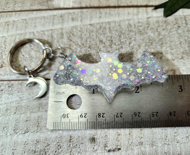 Pode incluir: Um chaveiro de morcego de glitter branco com um charme de lua crescente prateado. O morcego tem aproximadamente 5 cm de largura e est&aacute; preso a um chaveiro prateado.
