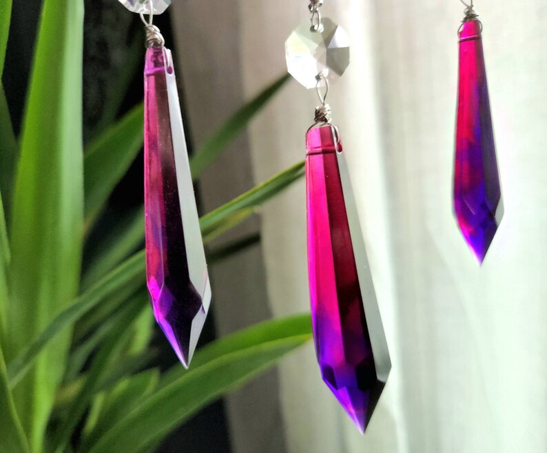 May include: Three crystal teardrop prisms with a gradient of pink and purple colors. The prisms are hanging from a silver wire.