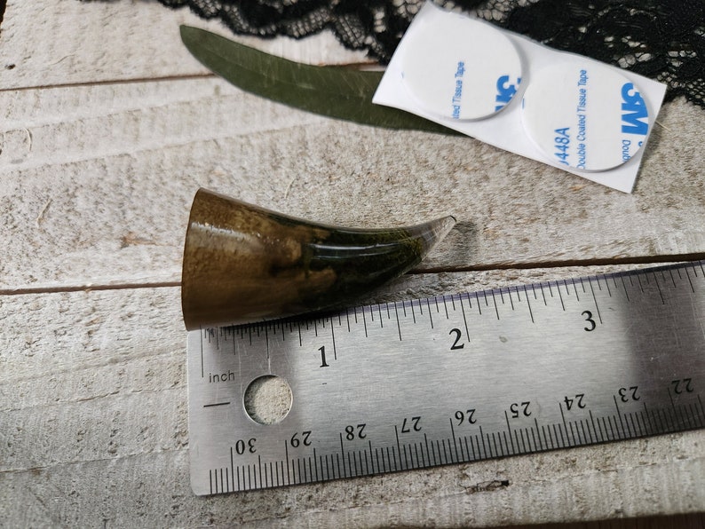 May include: A dark brown and green resin horn with a clear tip, measuring approximately 1 inch in length. The horn is sitting on a wooden surface next to a ruler.