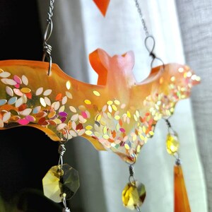 May include: Orange resin bat with iridescent glitter and crystal accents hanging from a silver chain. The bat is a Halloween decoration.