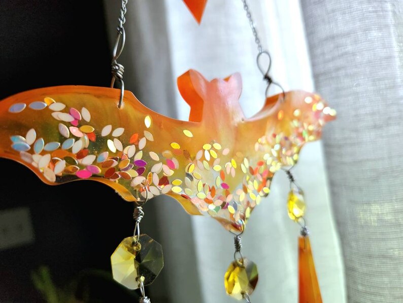 May include: Orange resin bat with iridescent glitter and crystal accents hanging from a silver chain. The bat is a Halloween decoration.
