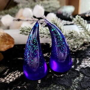 May include: A pair of purple resin horns with iridescent green glitter embedded in the resin. The horns are pointed and have a slightly curved shape.