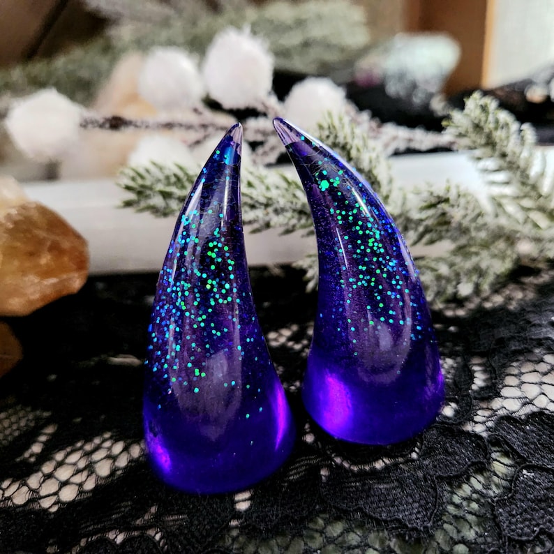 May include: A pair of purple resin horns with iridescent green glitter embedded in the resin. The horns are pointed and have a slightly curved shape.