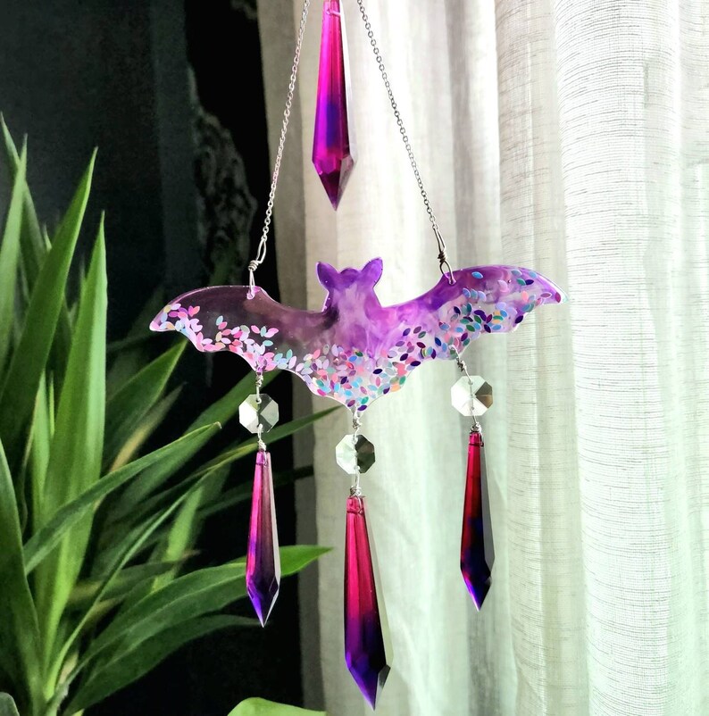 May include: A purple and pink bat-shaped resin ornament with iridescent glitter and crystal accents hanging from a silver chain.