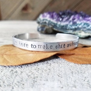 May include: A silver cuff bracelet with the stamped phrase "I'm here to make shit awkward." The bracelet is resting on a textured surface with a dried leaf and a purple crystal in the background. The bracelet is a simple, yet bold accessory.